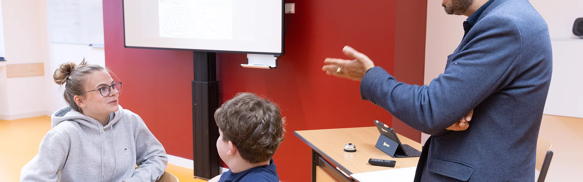 Lehrer mit dunkel braunen kurzen Haaren trägt eine Brille und ein blaues Hemd. Er hat die Arme verschränkt und schaut lächelnd auf die Schülerinnen und Schüler, welche ihre Finger in die Luft heben.Im Hintergrund eine grüne Kreidetafel mit einem dunkel roten Schwamm und einem Abzieher.