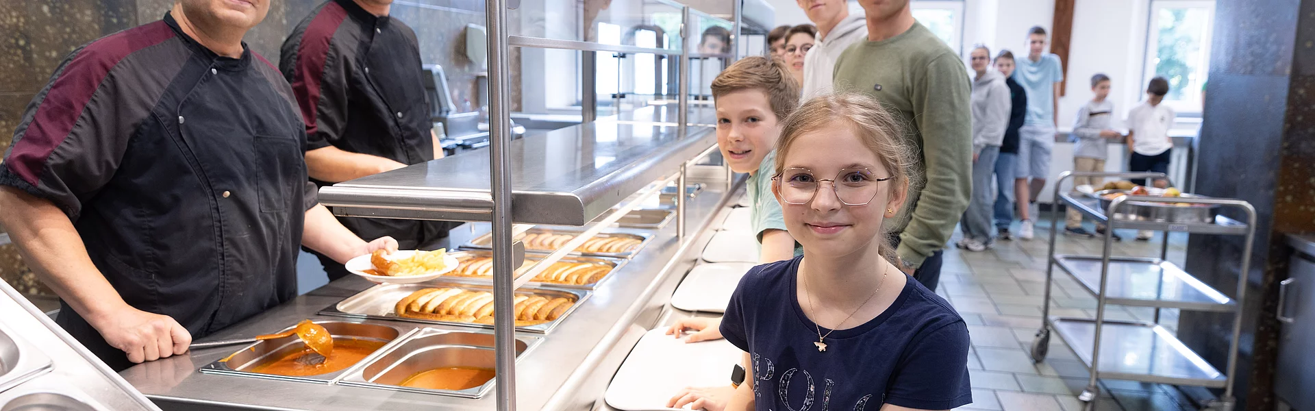Im Hintergrund stehen Schülerinnen und Schüler an der Essensausgabe. Im Vordergrund reicht ein Mann einem Schüler einen Teller mit einem Gericht.