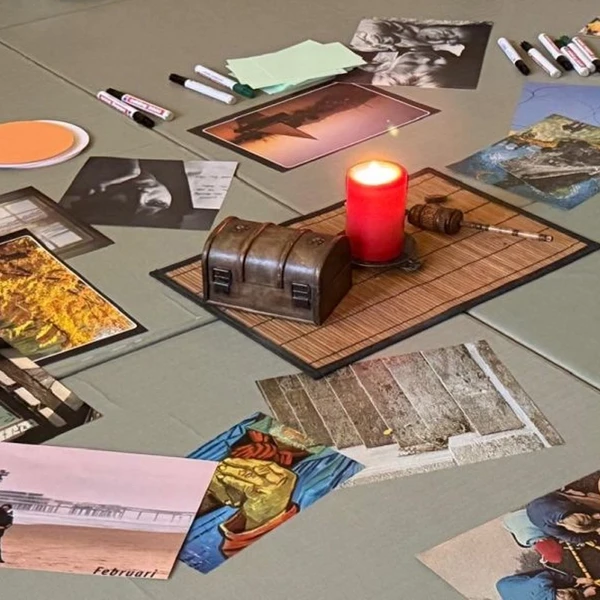 Auf dem Boden liegen eineige ausgedruckte Bilder verstreut, sowie bunte Stapel mit Blättern und verschiedene Eddings. In der Mitte liegt eine Bambusmatte, worauf eine rote leuchtende Kerze steht. Links neben der Kerze steht eine kleine dunkel-braune Truhe.