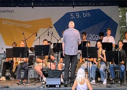 Schülerinnen und Schüler sitzen auf Stühlen auf einer Bühne und haben unterschiedliche Instrumente in der Hand. Auf der Bühne steht vor ihnen ein Mann.