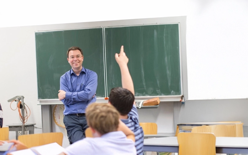 Lehrer mit dunkel braunen kurzen Haaren trägt eine Brille und ein blaues Hemd. Er hat die Arme verschränkt und schaut lächelnd auf die Schülerinnen und Schüler, welche ihre Finger in die Luft heben.Im Hintergrund eine grüne Kreidetafel mit einem dunkel roten Schwamm und einem Abzieher.